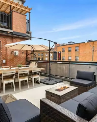 Rooftop terrace with dining table, lounge seating, fire pit, and umbrella overlooking brick buildings