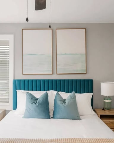 Bedroom with teal upholstered headboard, white bedding, blue pillows, wooden nightstands, and framed coastal artwork above bed