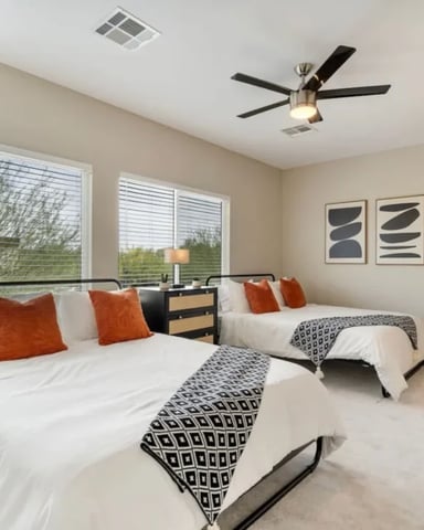 Bright bedroom with two queen beds, orange pillows, ceiling fan, large windows, and abstract wall art