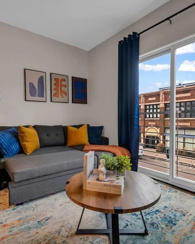 Modern living room with gray sofa, blue and yellow pillows, round wood coffee table, and floor-to-ceiling windows overlooking brick building and balcony.
