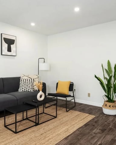 Modern living room with dark sofa, black metal tables, yellow accent chair, potted plant, and abstract wall art