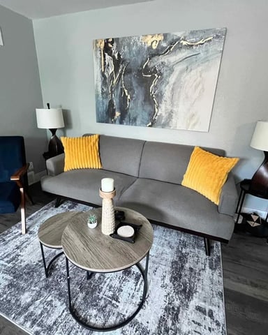 Modern living room with gray sofa, blue accent chair, round wood coffee table, yellow pillows, and abstract wall art