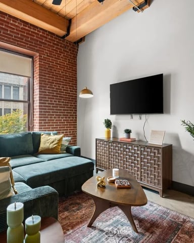 Modern loft living room with exposed brick wall, blue sectional sofa, wooden coffee table, wall-mounted TV, and large window overlooking city street.