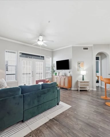 Open-plan living room with teal sofa, wall-mounted TV, kitchen island, and glass doors to balcony