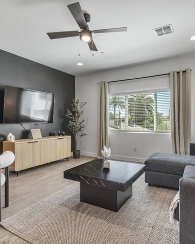 Modern living room with gray sectional sofa, black coffee table, wall-mounted TV, wood media console, and large windows with palm tree views.