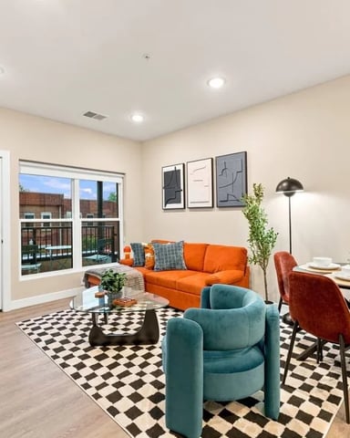 Open-plan living and dining area with orange sofa, teal armchair, black-and-white checkered rug, and glass doors opening to balcony with city views
