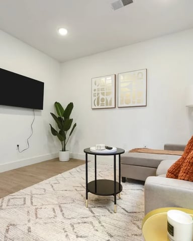 Modern bedroom with wall-mounted TV, gray upholstered bed with orange pillows, black side table, potted plant, and geometric wall art
