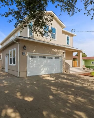 Beige two-story house with white garage, front porch, and driveway shaded by large tree.