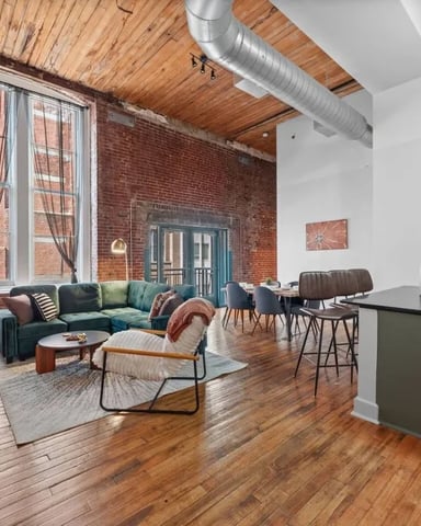 Industrial loft with exposed brick, wooden ceiling beams, green sectional sofa, and large windows overlooking city street.