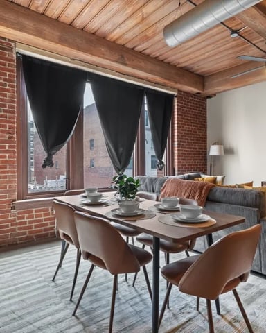 Loft dining area with exposed brick, wood ceiling, large windows with black curtains, and set dining table overlooking city buildings