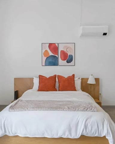 Bedroom with wood-frame bed, white bedding, orange pillows, abstract wall art, and wall-mounted air conditioning unit