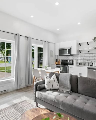 Open-plan living room and kitchen with gray sofa, white subway tile backsplash, stainless steel appliances, and glass doors opening to yard.