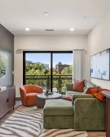Modern living room with green sectional sofa, orange accent chair, striped rug, and black-framed glass doors opening to city balcony view.