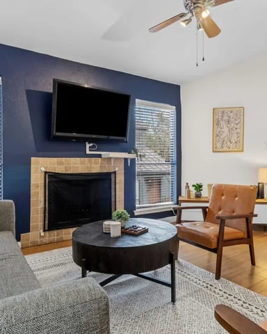 Living room with navy accent wall, fireplace, wall-mounted TV, round black coffee table, gray and cognac mid-century chairs, and wood flooring.