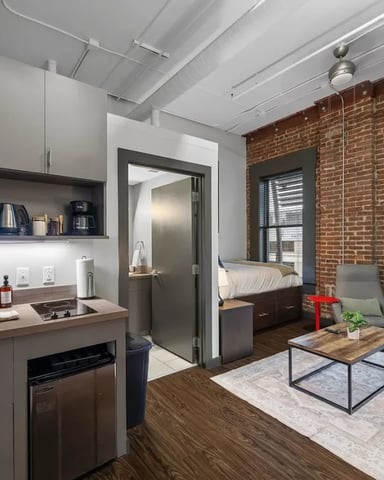 Modern studio apartment with exposed brick wall, kitchenette, living area with coffee table, and bedroom visible through doorway.