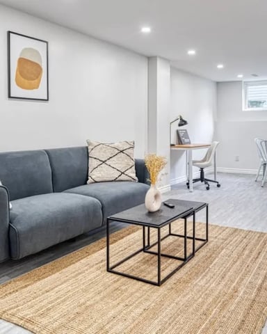 Finished basement with gray sofa, black coffee table, dining area, and desk under bright recessed lighting