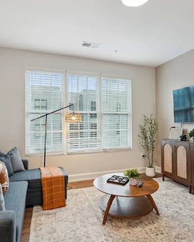 Modern living room with blue sofa, wooden coffee table, wall-mounted TV, and large windows overlooking city buildings