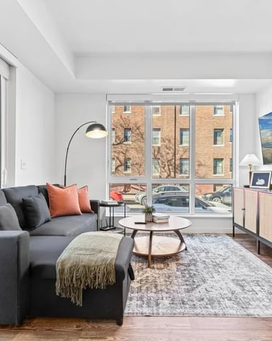 Modern living room with gray sofa, round coffee table, and large windows overlooking brick buildings and street parking