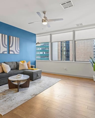 Modern living room with blue accent wall, gray sectional sofa, wood coffee table, and city views through large windows