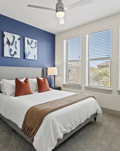 Bedroom with navy blue accent wall, gray upholstered bed, white bedding, rust pillows, multiple windows with blinds, ceiling fan, and potted plant on