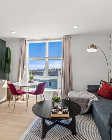 Modern living room with gray sofa, red pillows, black coffee table, green accent wall, dining area, and city skyline view through large windows