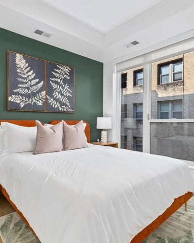 Bedroom with green accent wall, wood bed frame, white bedding, wooden nightstand with lamp, and large windows overlooking brick building.