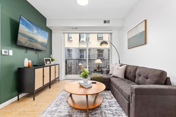 Modern living room with green accent wall, gray sofa, round wood table, and glass doors opening to balcony overlooking city street.