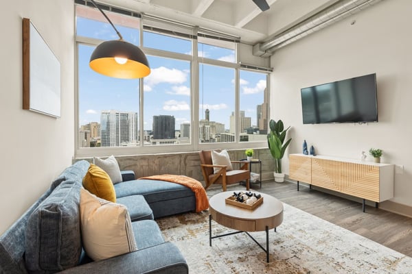 Modern high-rise living room with blue sectional sofa, city skyline view through floor-to-ceiling windows, and mid-century furniture.