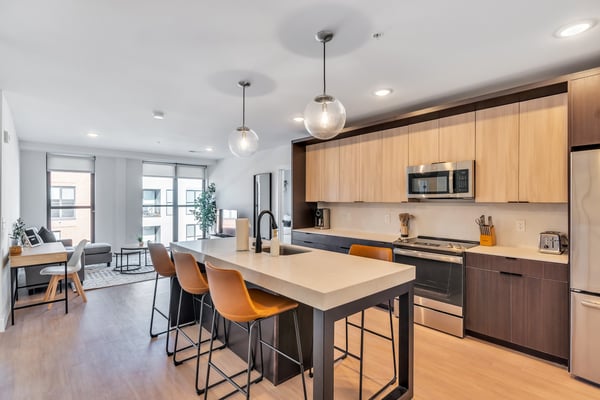 Modern open-plan kitchen and living room with light wood cabinetry, island seating, stainless steel appliances, and pendant lighting.