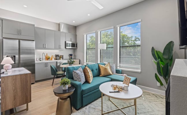 Modern open-plan living room with teal sofa, white oval coffee table, and integrated kitchen with stainless steel appliances and large windows with blinds.