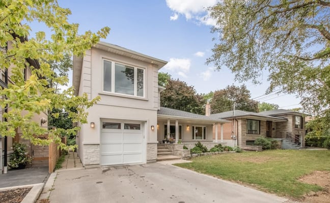 Modern two-story house with white garage, brick and stone exterior, front lawn, and mature trees overhead.