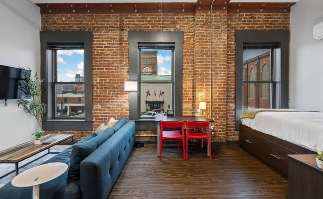 Studio apartment with exposed brick wall, blue sofa, red chairs, bed, and three windows overlooking urban street