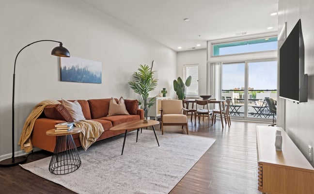 Modern living room with rust-colored sofa, arc lamp, potted plants, and floor-to-ceiling windows overlooking a balcony with city views