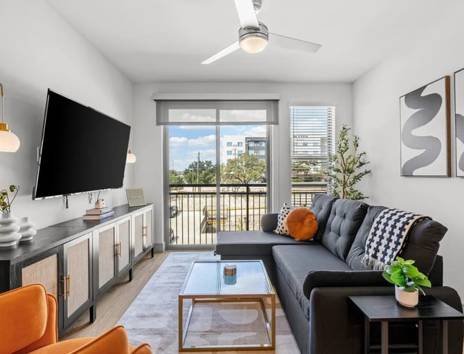 Modern living room with dark sectional sofa, wall-mounted TV, glass balcony doors overlooking city skyline, and contemporary furnishings.